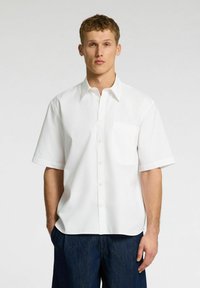 Camicia bianca a maniche corte realizzata in tessuto liscio, con colletto a portafoglio e taschino sul petto. Abbinata a pantaloni in denim blu scuro.