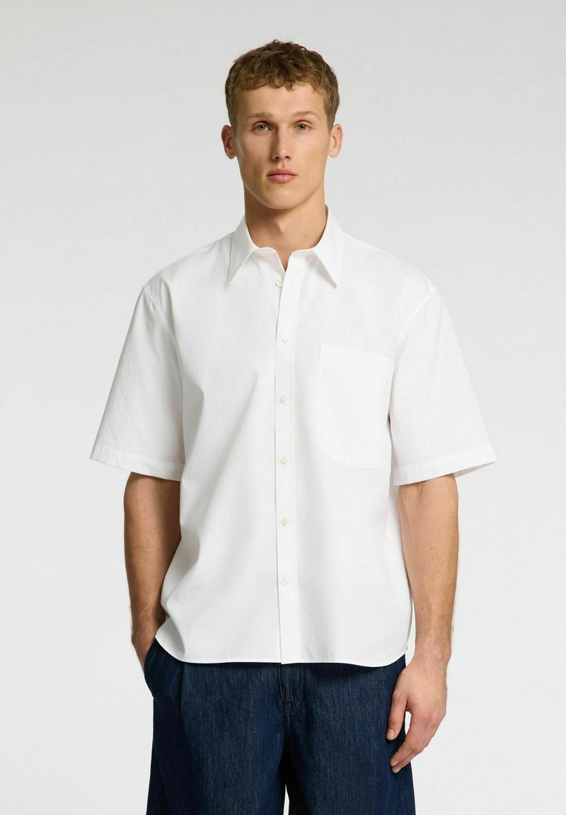 Camicia bianca a maniche corte realizzata in tessuto liscio, con colletto a portafoglio e taschino sul petto. Abbinata a pantaloni in denim blu scuro.