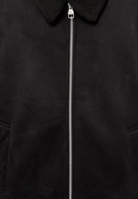 Veste noire à texture douce avec une fermeture éclair centrale argentée et un col en polaire noir, avec deux poches latérales partiellement visibles.