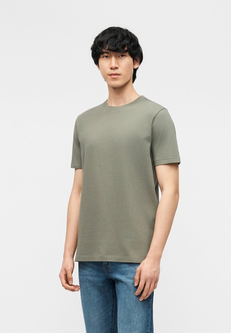 Νέος άνδρας φοράει ένα απλό ελιάς κοντομάνικο μπλουζάκι και μπλε τζιν, στέκεται μπροστά σε ανοιχτό γκρι φόντο.