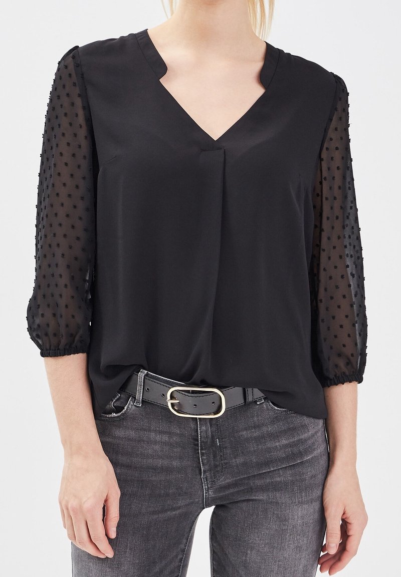 Femme portant un chemisier noir à manches trois-quarts transparentes à pois, associé à un jean gris foncé et une ceinture noire avec une boucle dorée.
