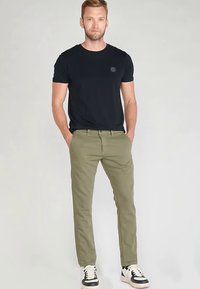 Camiseta negra de manga corta con un pequeño logo, combinada con pantalones chinos verde oliva claro y zapatillas negras y blancas. Fondo liso.