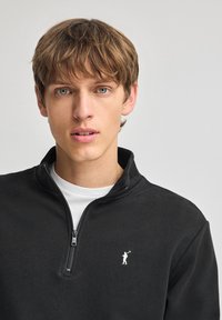 Zwarte zip-up sweatshirt met een hoge kraag, voorzien van een klein wit golferlogo op de borst. Gemaakt van een zachte, gestructureerde stof.