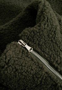 Σκούρο πράσινο φλις με υφή στην επιφάνεια που διαθέτει ασημί φερμουάρ. Το φερμουάρ εκτείνεται κατά μήκος μιας ραφής, αναδεικνύοντας την απαλότητα του υλικού.
