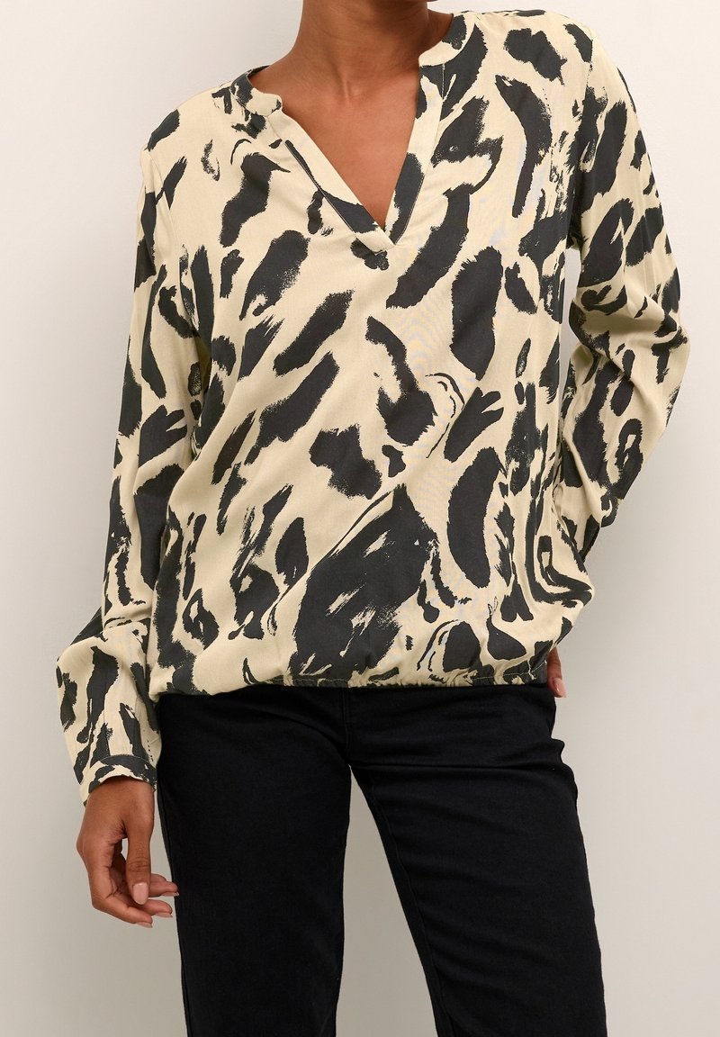 Femme portant une blouse beige avec des taches abstraites noires et un pantalon noir, posant avec une main dans la poche.