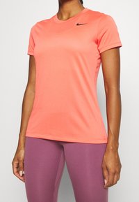 Mujer vestida con una camiseta atlética de manga corta en coral brillante con el logo negro de Nike y leggings ajustados morados, de pie contra un fondo claro y liso.