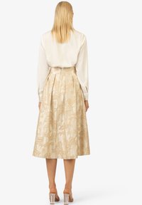 Jupe beige avec broderie florale, design plissé et ceinture. Portée avec un blouse blanche à manches longues et des sandales à talons transparents.