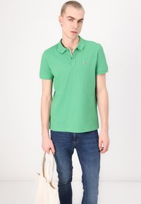 Grønn bomullspolo med krage, korte ermer og en liten logo på brystet; kombinert med blå denimjeans og en lerretstotebag.