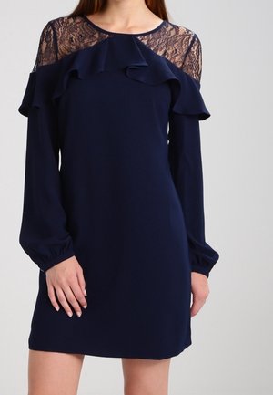 Femme portant une robe à manches longues bleu marine avec des épaules en dentelle et un détail volanté sur la poitrine et le haut des bras.