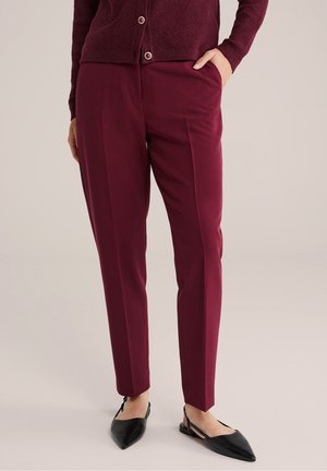 Vrouw draagt bordeauxrode, op maat gemaakte broek en bijpassend textuurvest met zwarte, puntige slingback ballerina's, staand met één hand in de zak.