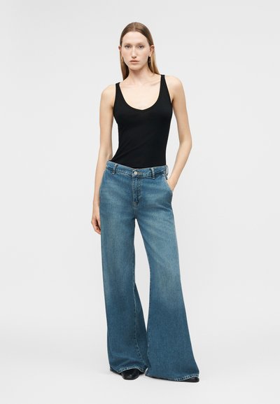 Νεαρή γυναίκα φορά μαύρο αμάνικο τοπ και μπλε τζιν με ψηλή μέση και φαρδιά μπατζάκια, στέκεται με το ένα χέρι στην τσέπη μπροστά σε μονόχρωμο φόντο.