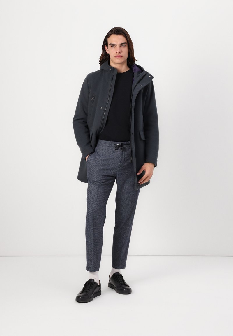 Giacca grigia scura con cappuccio, maglietta nera e pantaloni blu a motivo. Scarpe da ginnastica nere con calze bianche. Design semplice, look casual.