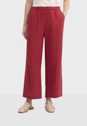 Czerwone lniane spodnie o szerokich nogawkach z elastycznym pasem i bocznymi kieszeniami, z teksturowanym wykończeniem i luźnym krojem. Noszone z jasnymi sandałami.