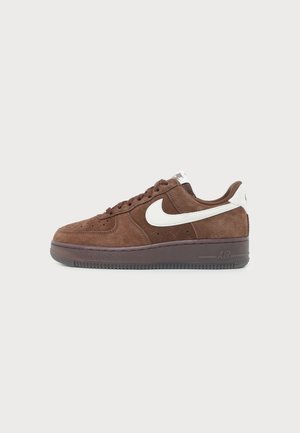 Niski, brązowy zamszowy sneaker z białym logo Nike, sznurowadłami i ciemnobrązową podeszwą na jednolitym jasnoszarym tle.