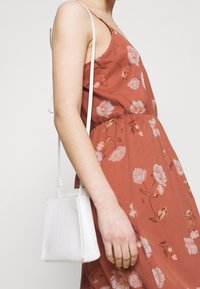 Abito color ruggine con motivo floreale; tessuto leggero e vestibilità ampia. Piccola borsa bianca con una finitura testurizzata, indossata su una cinturina sottile.
