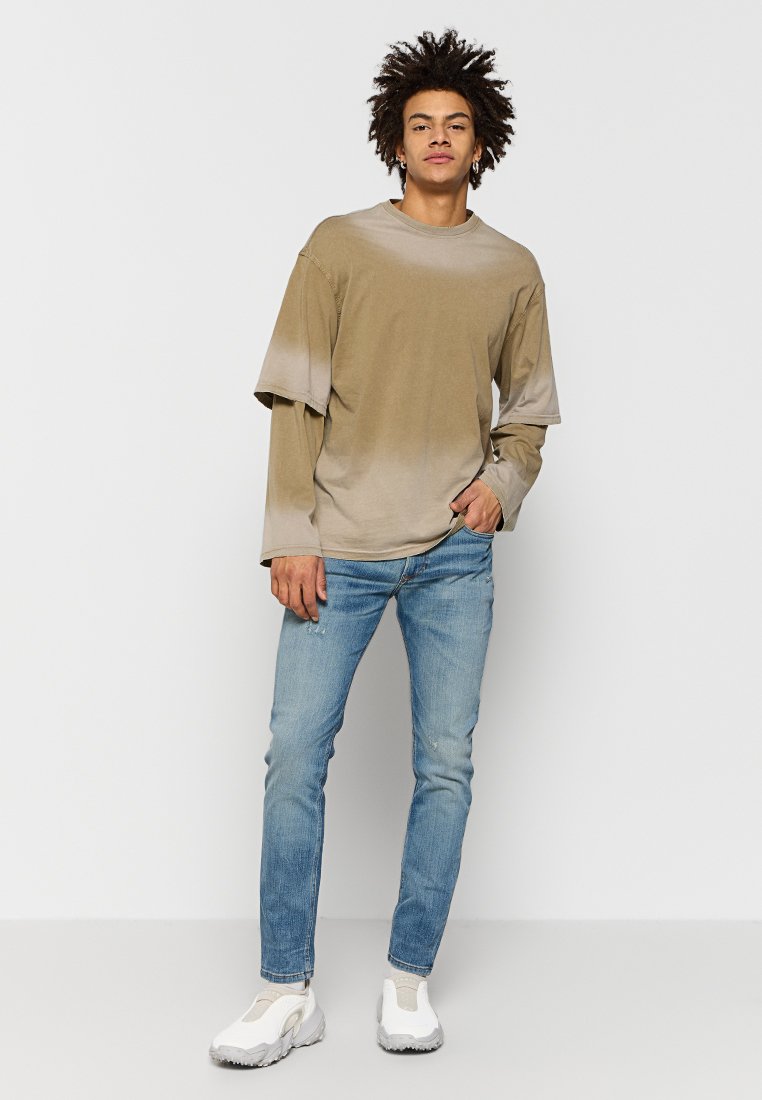 Jeune homme avec des cheveux volumineux portant des chemises à manches longues et courtes beiges superposées, un jean bleu clair et des baskets blanches se tenant contre un fond blanc.