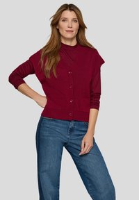 Bordeauxroter Cardigan mit Knopfleiste, kurzen Ärmeln und zwei Fronttaschen. Getragen über einem passenden roten Oberteil und kombiniert mit blauen Jeans.