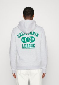 Persona con felpa grigia chiara con la scritta "California League 1774 College Championship" e grafica di baseball sulla schiena, che guarda di spalle.