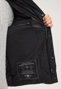 Giacca nera con una texture liscia, dotata di una tasca interna con zip e un’etichetta che indica che è imbottita con il 100% di poliestere riciclato.