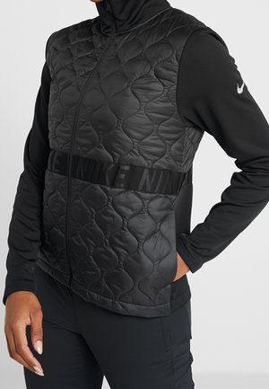 Person trägt schwarze, gesteppte Nike-Weste über einem langärmeligen schwarzen Oberteil, mit der Hand in der Tasche, dargestellt vom Hals bis zum Oberschenkel vor neutralem Hintergrund.