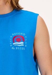 Mėlyna be rankovių medvilninė marškinėlių palaidinė su siuvinėtu logotipu, kuriame pavaizduota raudona saulė ir mėlynos bangos, ir užrašu "BEACH BEYOND CLUB the BLUES."
