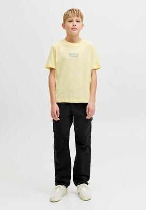 Lyshåret dreng står med fronten mod kameraet iført en bleg gul T-shirt med tryk, sorte cargobukser og hvide sneakers mod en ensfarvet baggrund.