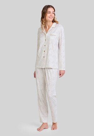 Una mujer de pie descalza, vistiendo un pijama blanco de manga larga con botones, adornado con pequeños lunares azules y negros, sobre un fondo gris liso.