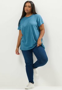 Sininen ribattu lyhythihainen paita, jossa on löysä malli, yhdistetty tummansinisiin kapeisiin farkkuihin ja valkoisiin lenkkareihin neutraalilla taustalla.