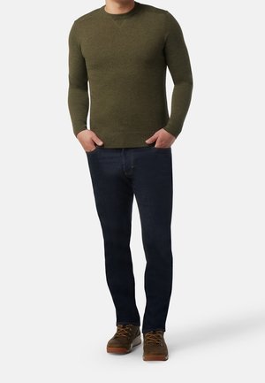 Homme portant un pull à manches longues vert olive, un jean bleu foncé et des chaussures de randonnée brunes, se tenant avec les mains dans les poches sur fond blanc.