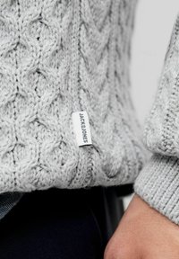 Grauer Strickpullover mit einem Kabelmuster, strukturiertem Finish und einem kleinen Etikett mit dem Markennamen auf der unteren Seite.