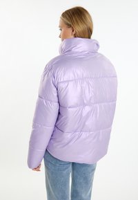 Giacca puffer imbottita in lavanda chiaro, dotata di un colletto alto, design trapuntato e finitura lucida. Indossata con jeans blu casual.