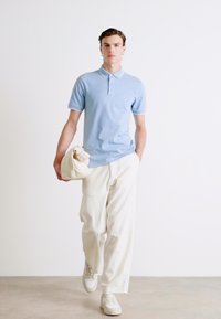 Polo shirt azzurro chiaro con rifiniture bianche, abbinato a pantaloni bianchi larghi. Il modello tiene una borsa crema, indossando sneaker bianche. Tessuto morbido, design casual.