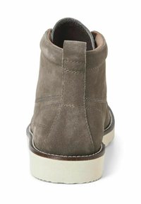 Stivale alla caviglia in suede marrone con punta arrotondata, dettagli cuciti, fodera color sabbia e suola in gomma bianca. Presenta una linguetta sul retro.