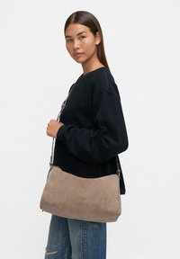Borsa a tracolla in suede di un color taupe chiaro, con forma arrotondata, hardware in argento e tracolla regolabile. Superficie testurizzata e branding minimalista.