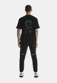 Fekete póló, hátán textúrált grafikai dizájnnal; fekete nadrággal párosítva, amely visszaverő elemekkel és karcsúsított fazonnal rendelkezik.