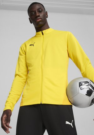 Geel sportjack met een rits aan de voorkant en een hoge kraag. Zwart logo op de linkerborst. Model houdt een voetbal vast en draagt zwarte broek.