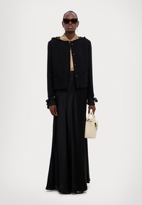 Cardigan negru cu textură, cu margini destrămate, asortat cu o fustă lungă de satin negru. Ține în mână o geantă crem, având mâneci scurte și fundițe accent.