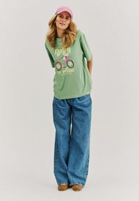 Woman wearing a pink cap, green oversized t-shirt with bicycle graphic, wide blue jeans, and brown shoes, standing against a plain background.