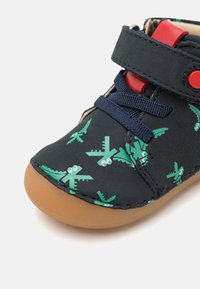 Chaussure en tissu bleu avec des motifs d'avion verts, des accents rouges et une semelle en caoutchouc. Possède une sangle en velcro et des lacets navy pour un maintien sécurisé.