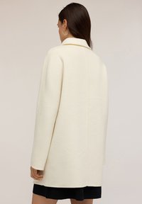 Cappotto in misto lana color crema con vestibilità rilassata, colletto sciallato e spalle basse. Presenta un design pulito e minimalista senza chiusure visibili.