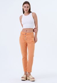 Un débardeur blanc coupé associé à un pantalon en coton orange avec une fermeture à bouton, des passants de ceinture et des accents cousus. Des sandales à plateforme complètent le look.