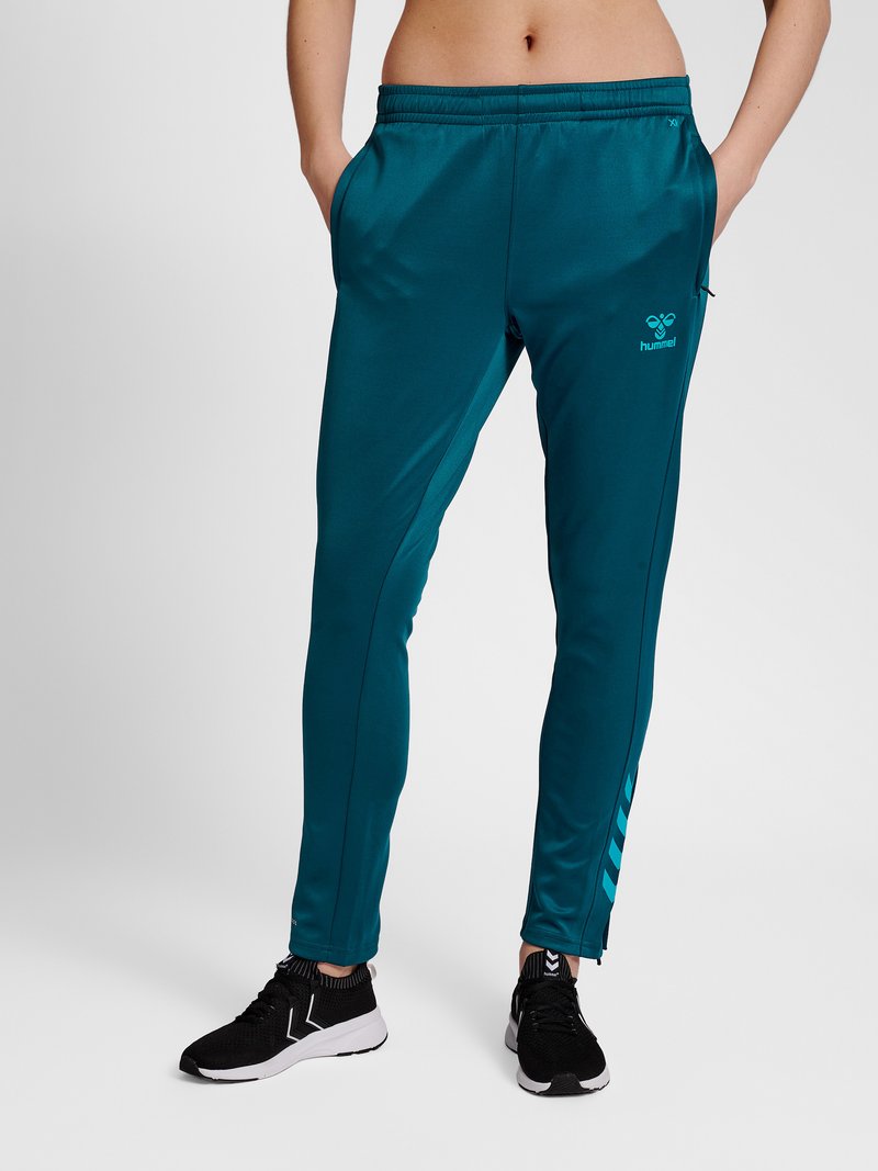 Persoon draagt turquoise sportbroek met zijzakken en zwarte sneakers, staat met handen in de zakken tegen een effen achtergrond.