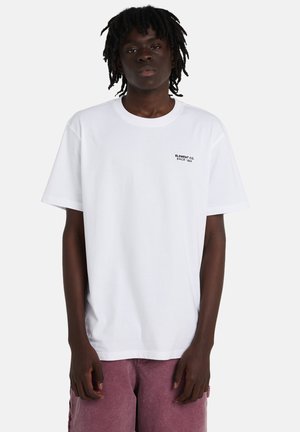 T-shirt en coton blanc à manches courtes, col rond, avec un petit logo texte noir sur la poitrine gauche. Porté avec un short rose coupe décontractée.