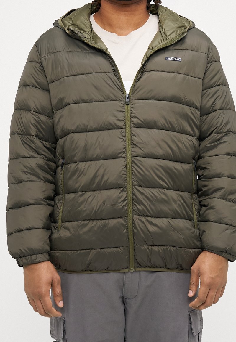 Homme portant une doudoune vert olive avec fermeture éclair et poches latérales, par-dessus une chemise blanche et un pantalon cargo gris.