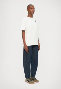T-shirt bianca in cotone con maniche corte e logo ricamato sul petto, abbinata a jeans in denim blu a vestibilità larga e sneakers verdi.