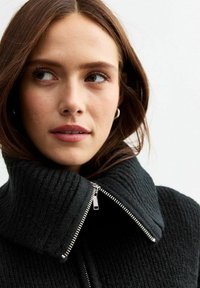 Svart texturerad jacka med hög krage och silverfärgad dragkedja. Har ribbat tyg och en närbild av halsringningen och kragdetaljerna.