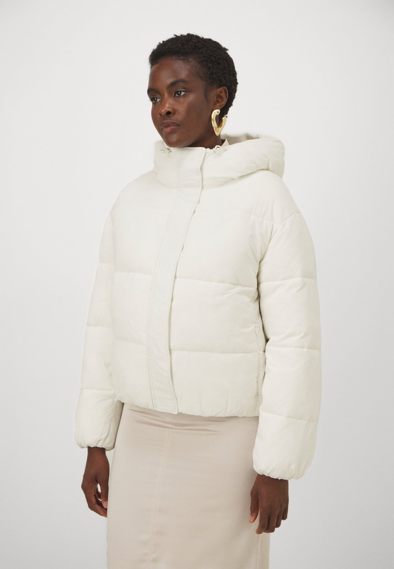 Giacca a vento in morbido tessuto bianco, con cappuccio, zip frontale e sezioni oversize trapuntate. Texture liscia con polsini elasticizzati.