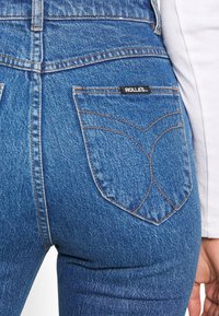 Džínové kalhoty s vysokým pasem v modré barvě, které mají výrazný zakřivený šev na zadních kapsách, a černou nášivku s nápisem "ROLLAS."