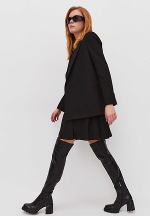 Sacou negru croitorit peste o fustă plisată neagră, asortat cu cizme negre lucioase până la coapsă, cu toc gros și fermoar lateral.