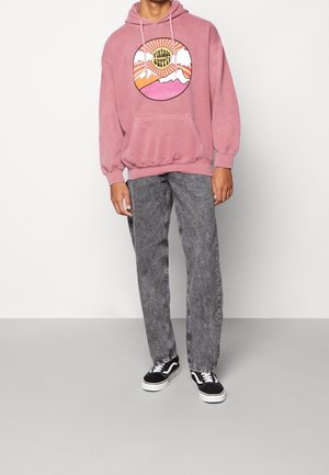 Rosa hoodie med en grafik av berg och sol. Gråa jeans med syrafärgning och svarta sneakers kompletterar outfiten. Avslappnad, ledig passform.